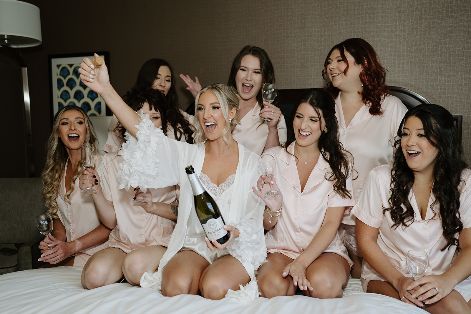 Bride and bridesmaids in pink pajamas cheer on a bed, with the bride holding a bottle of Prosecco.